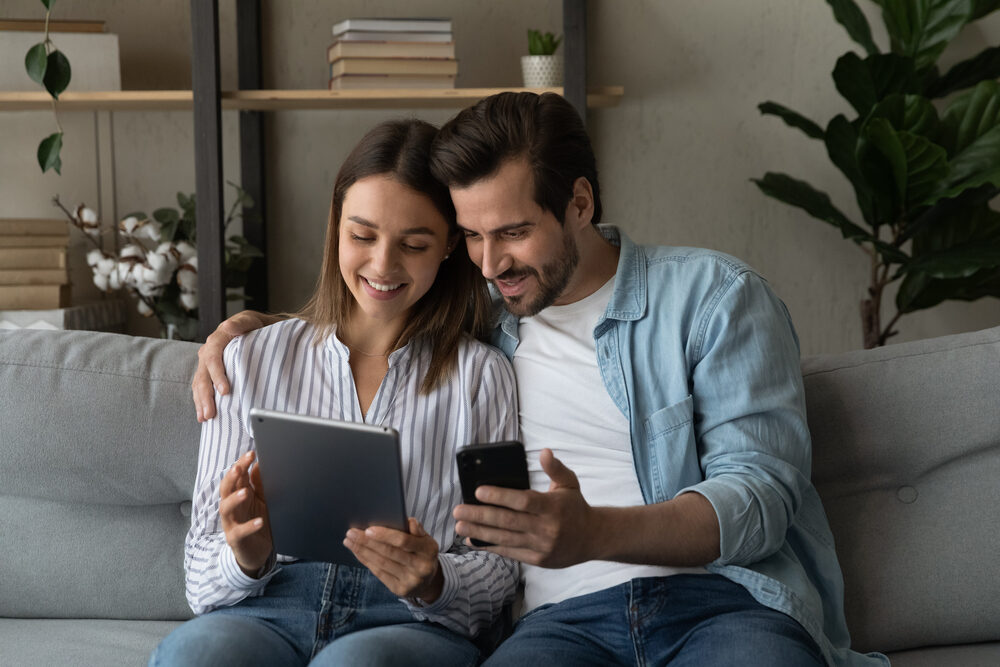 Couple Reviewing Home Selling Options Online – We Buy Houses Richland Smiling couple sitting on a couch using a tablet and phone to explore home selling options – We Buy Houses Richland