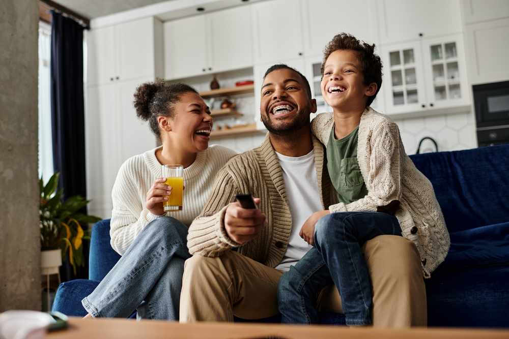 Smiling Family Relaxing at Home – Sell My House Fast Escondido Happy family sitting together on a couch in a modern living room, laughing and watching TV, symbolizing comfort and new beginnings after selling their home – Sell My House Fast Escondido