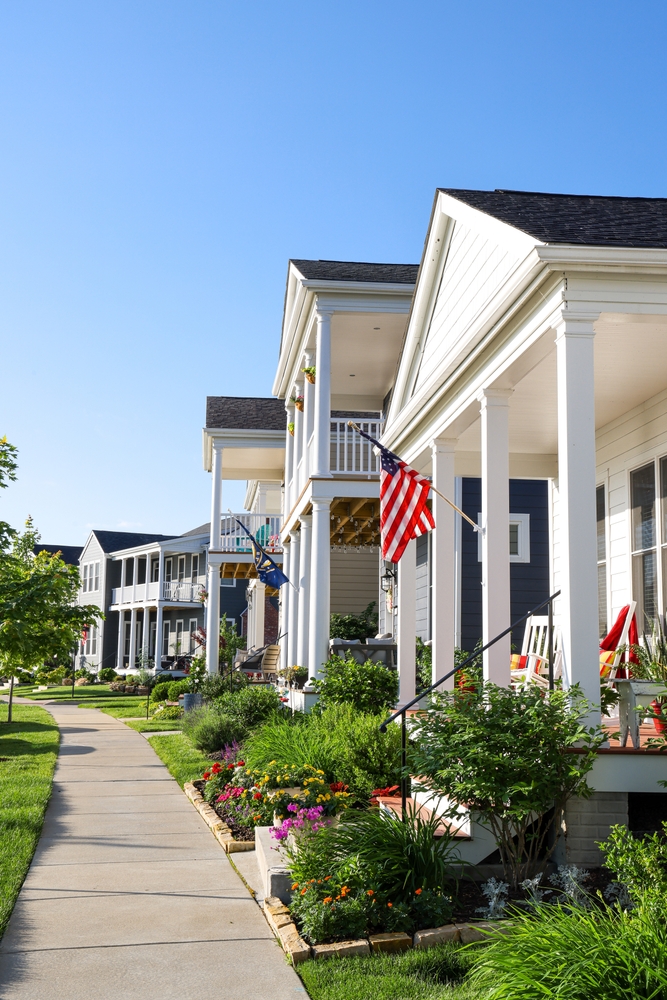 Beautiful Neighborhood Street with Modern Front Porches – JP Buys SD Row of well-kept homes featuring front porches, gardens, and American flags along a sunny suburban sidewalk – JP Buys SD