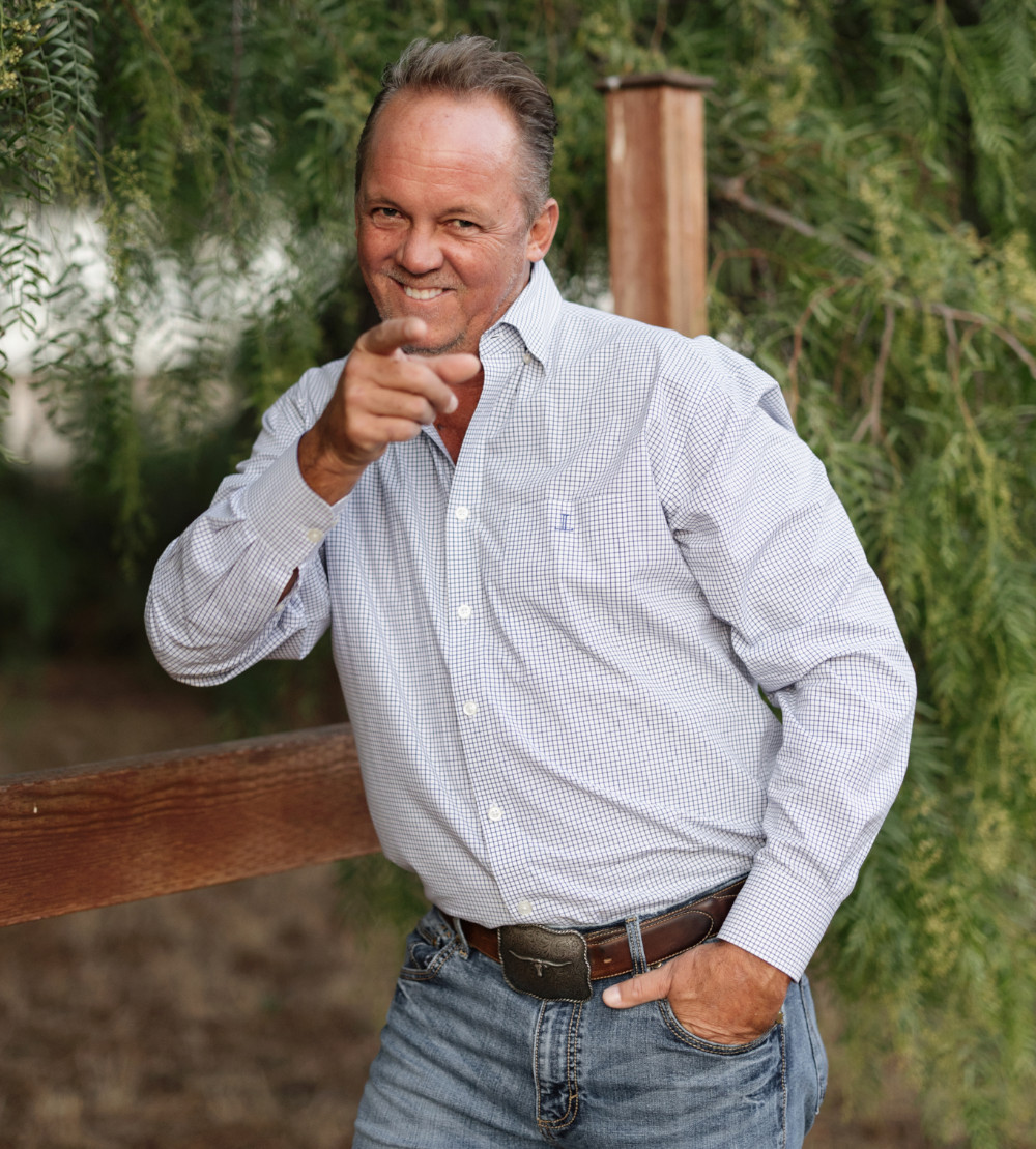 Jarrett Pugh Pointing Playfully Outdoors – JP Buys SD Jarrett Pugh in a button-down shirt and jeans pointing toward the camera while standing near a wooden fence and greenery – JP Buys SD