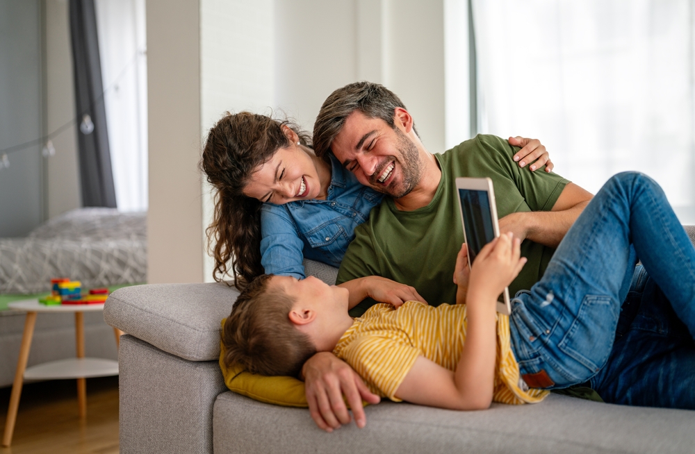 Happy family sitting on a sofa laughing together while their child holds a tablet, symbolizing comfort, joy, and a stress-free home-selling process – Sell My House Fast El Cajon