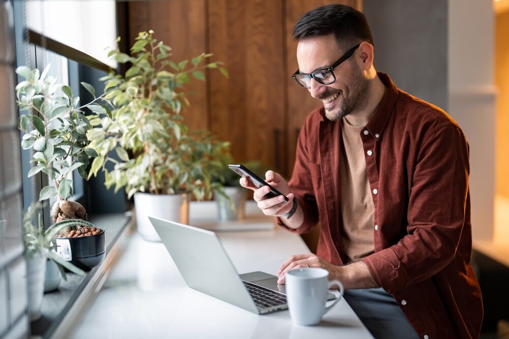 Man Working from Home on Laptop – Sell My House Oceanside Smiling man using laptop and smartphone near a sunny window, representing convenience in selling homes online – Sell My House Oceanside