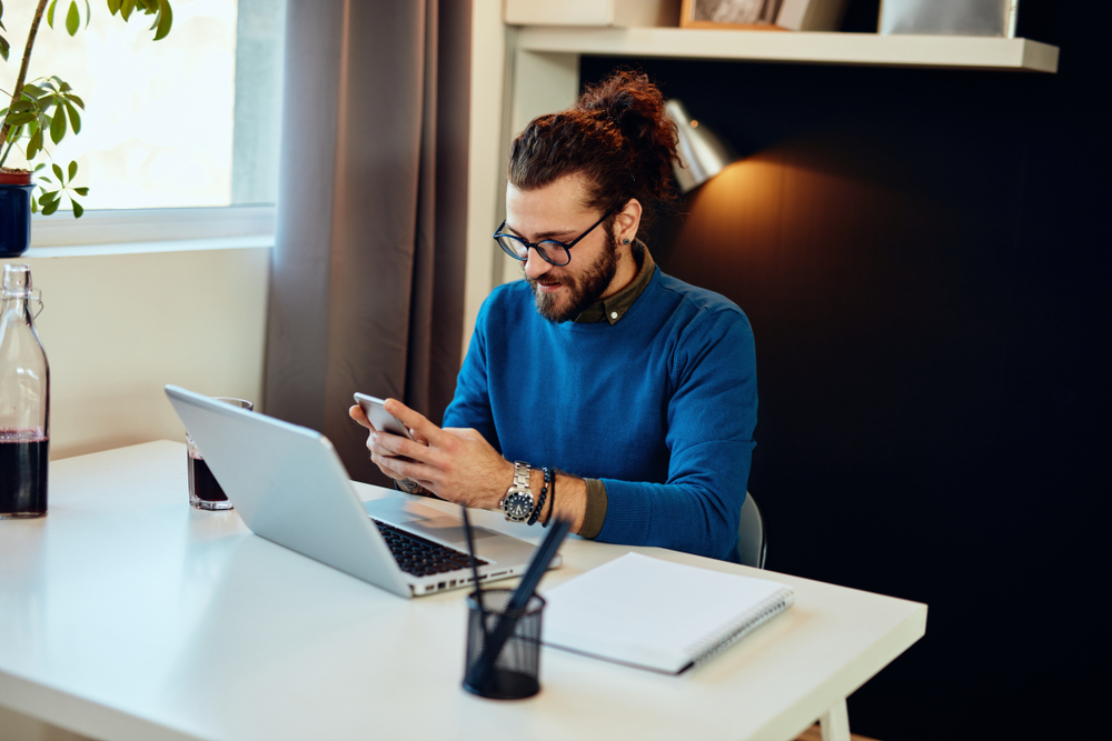 Young man sitting at a desk using his smartphone and laptop, checking property details online – We Buy Houses Chula Vista