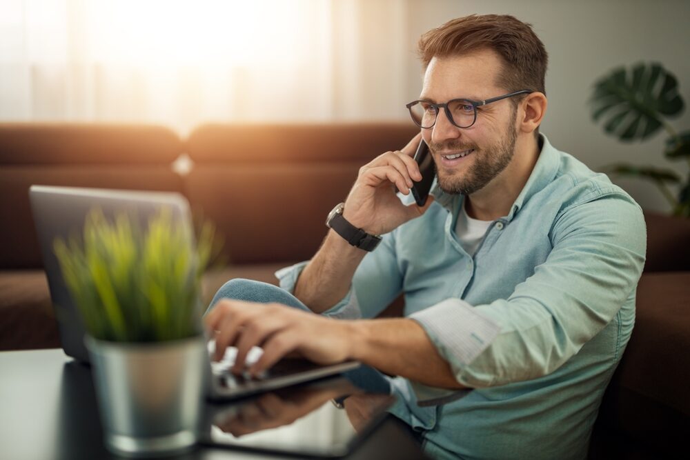 Professional Working Remotely – Property Appraiser San Diego CA Man with glasses talking on the phone and using a laptop at home, symbolizing professional real estate communication and property appraisal services – Property Appraiser San Diego CA