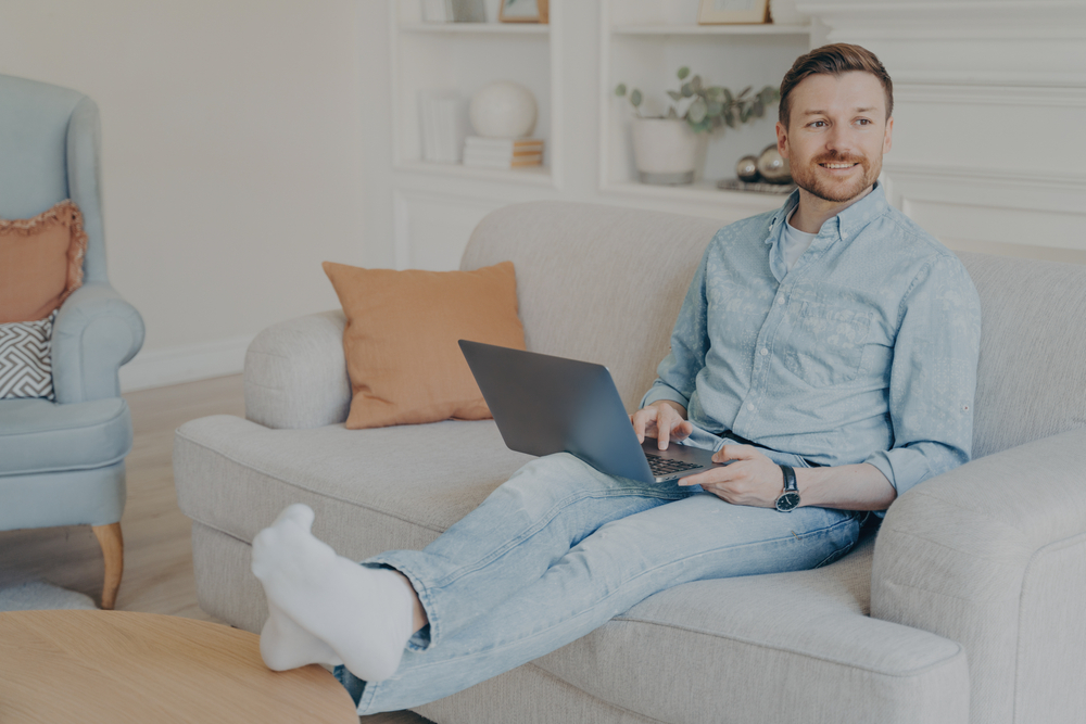 Man Relaxing on Couch While Working on Laptop – JP Buys SD Smiling man sitting comfortably on a sofa with a laptop on his lap in a bright living room – JP Buys SD