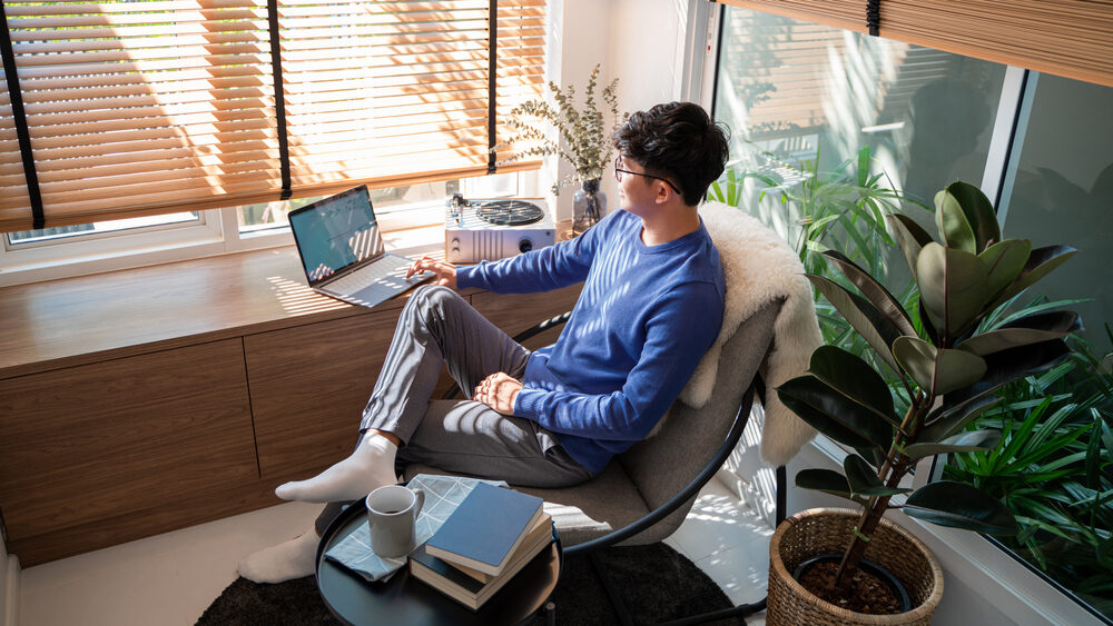 Man Working from Home by the Window – Sell My House Fast in San Marcos Young man sitting by a sunny window with a laptop, representing comfort and ease after a quick home sale – Sell My House Fast in San Marcos