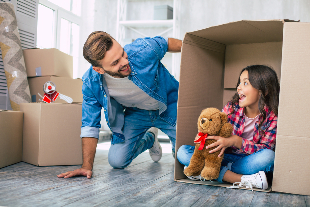 Smiling father kneeling beside his daughter playing with a teddy bear inside a moving box, symbolizing a happy family preparing for a smooth home sale and move – Sell My House Fast El Cajon