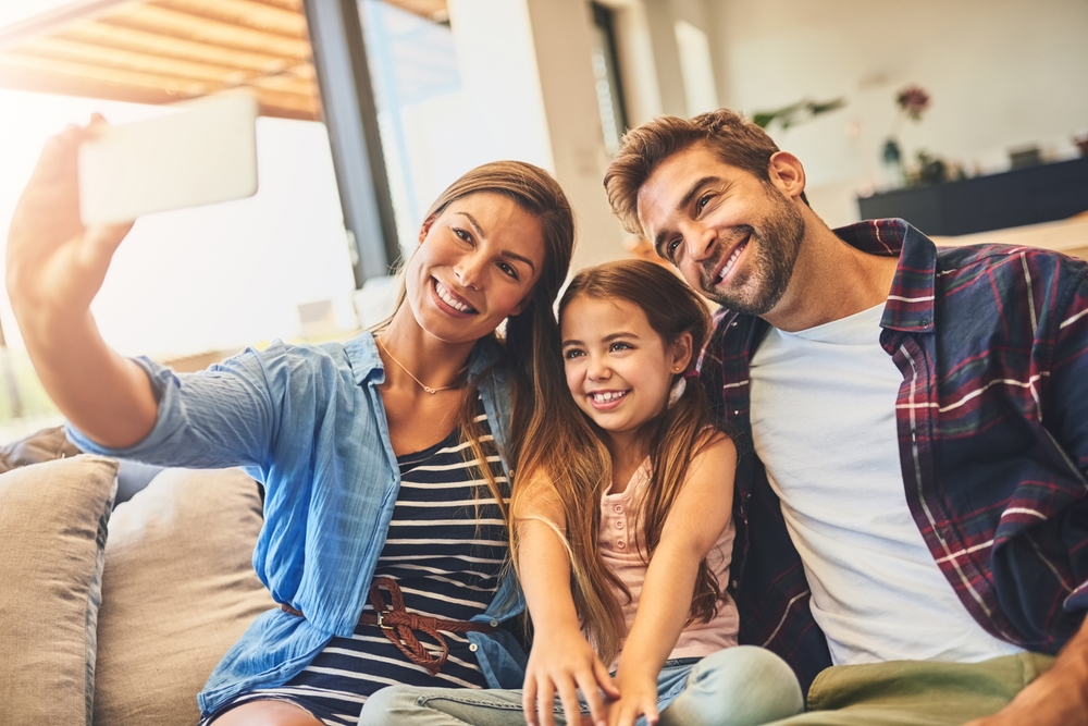 Smiling Family Taking a Selfie in Their New Living Room – Sell My House Oceanside Happy family sitting together on the couch taking a cheerful selfie after selling their home – Sell My House Oceanside