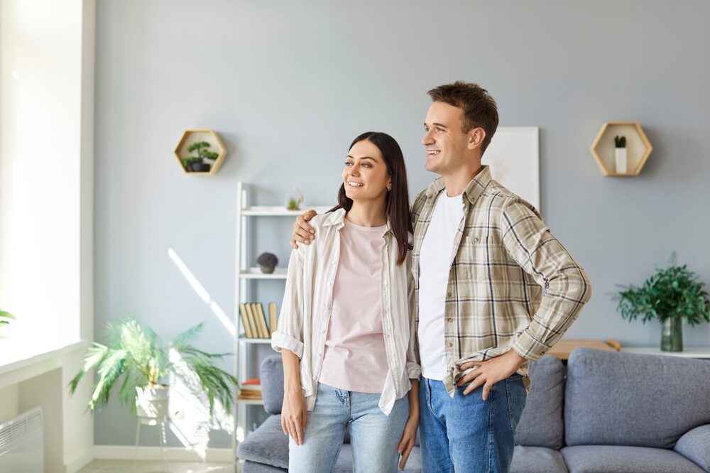 Smiling couple standing together in a bright modern living room, looking out the window with satisfaction after purchasing their dream home – Mission Valley Home Buyers