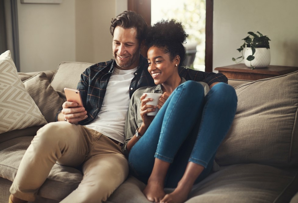 Happy Couple Relaxing at Home – Property Appraiser San Diego CA Smiling couple sitting together on a couch, looking at a smartphone and enjoying a relaxed moment at home, representing property ownership satisfaction – Property Appraiser San Diego CA
