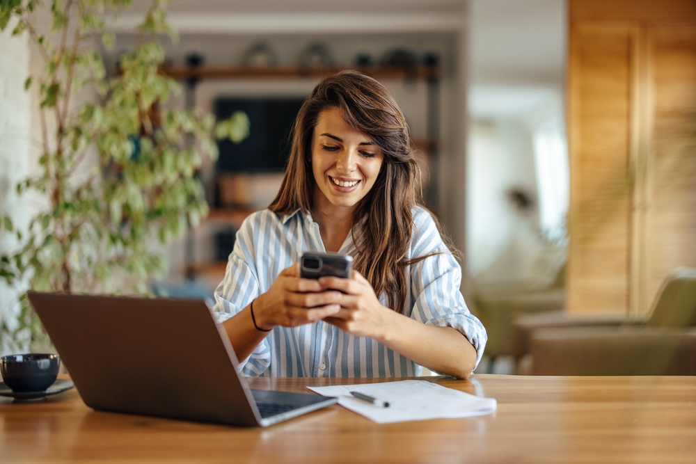 Smiling Woman Working from Home – we buy houses vista Smiling woman sitting at a desk using her phone beside a laptop and paperwork, representing professional communication and reliable property appraisal services – we buy houses vista