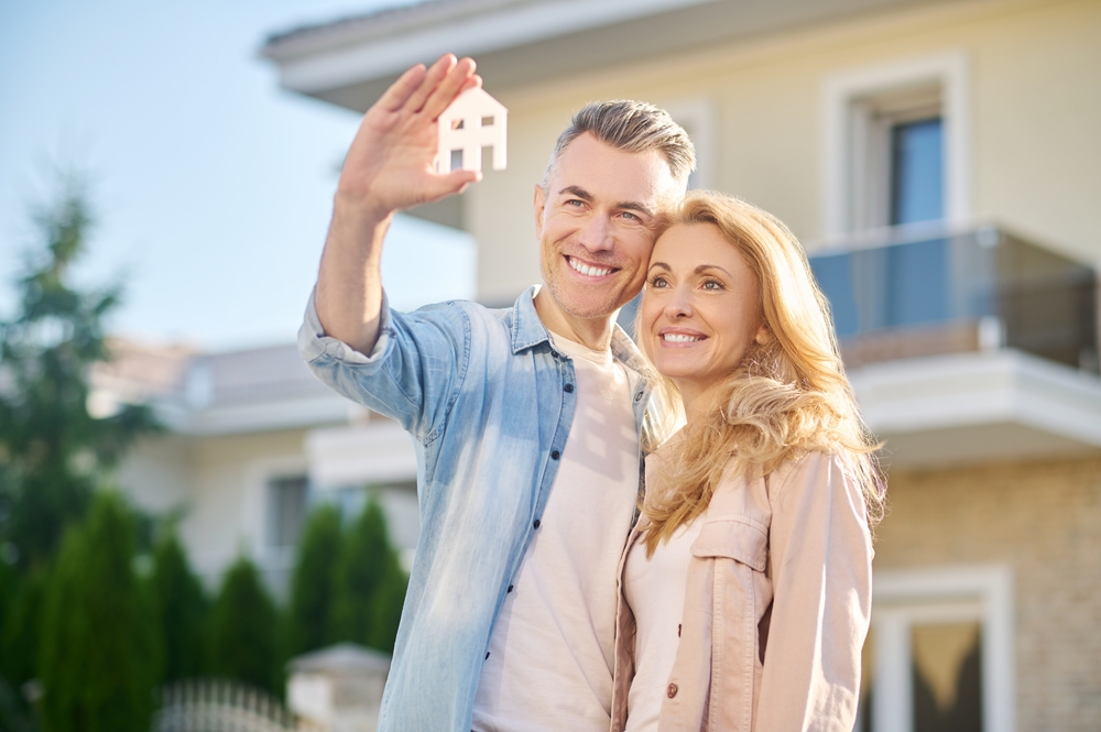 Happy Homeowners with New Property – Property Appraiser San Diego CA Smiling couple standing outside their new house while holding a small home model, representing accurate property valuation and real estate success – Property Appraiser San Diego CA