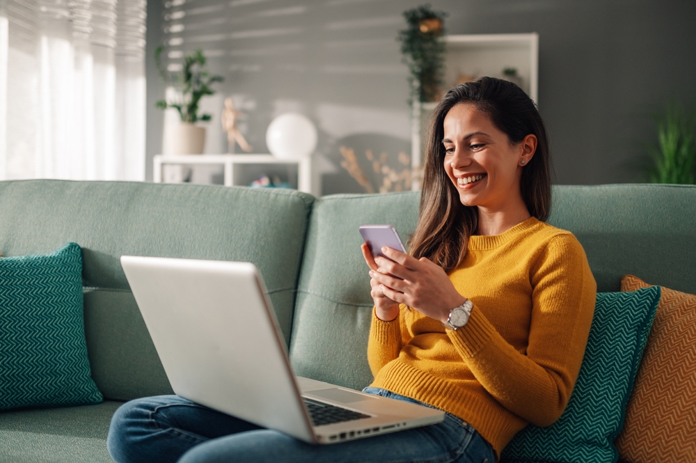 Smiling woman sitting on a couch using her smartphone while working on a laptop, symbolizing convenient communication and seamless home buying experience – Mission Valley Home Buyers